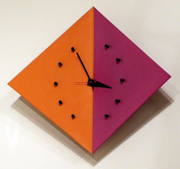 Kite Clock, en romb i orange och magenta med svarta pinnar som timmarkeringar och svarta visare, på en vit vägg