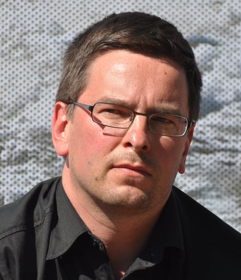 Harri Koskinen i bröstbild, med glasögon och svart skjorta, framför en ljus perforerad vägg