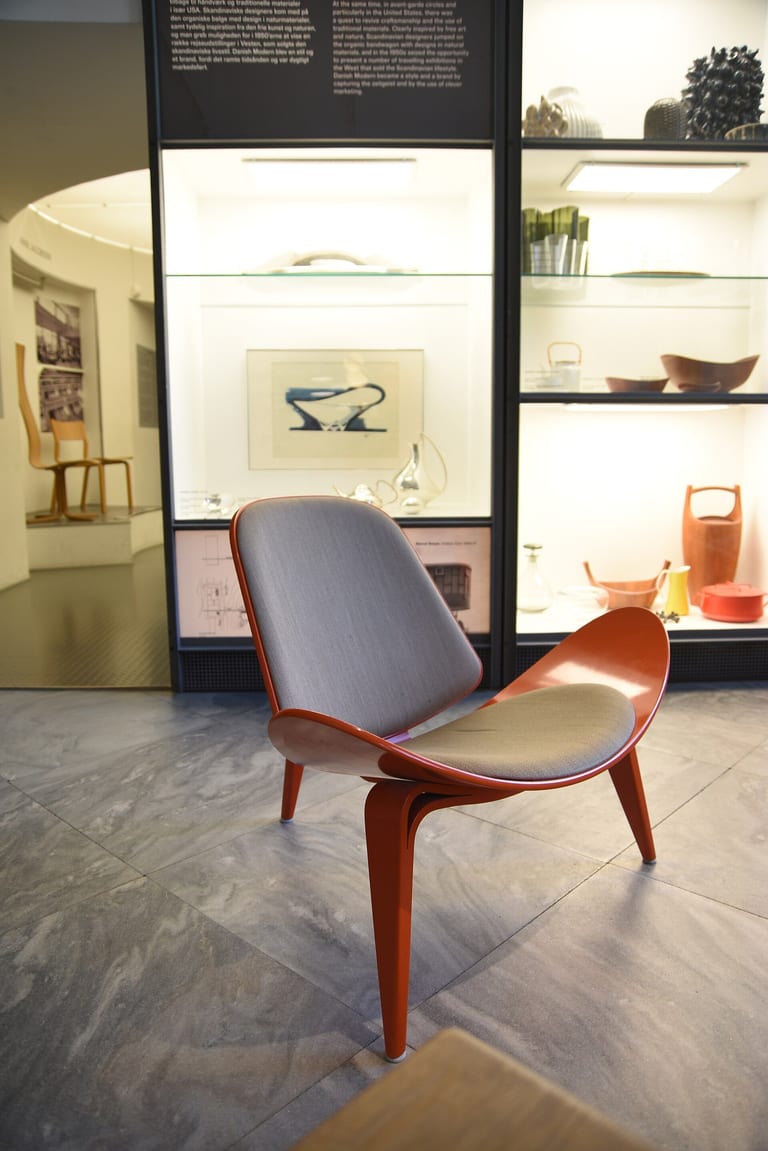 Shell Chair med röd stomme och grå klädsel, sedd snett från sidan i en museisal