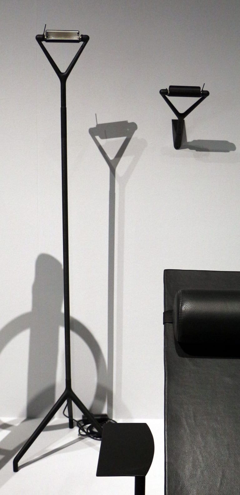 Svart golvlampa Lola på trebent fot med triangulärt huvud och en vägglampa Lola på en vit vägg, med en svart läderschäslong i förgrunden till höger