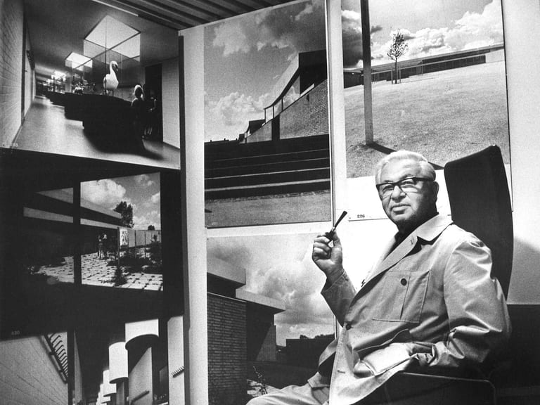 Svartvitt fotografi av Arne Jacobsen i glasögon och ljus rock, sittande med en pipa i handen framför stora fotografier av byggnader