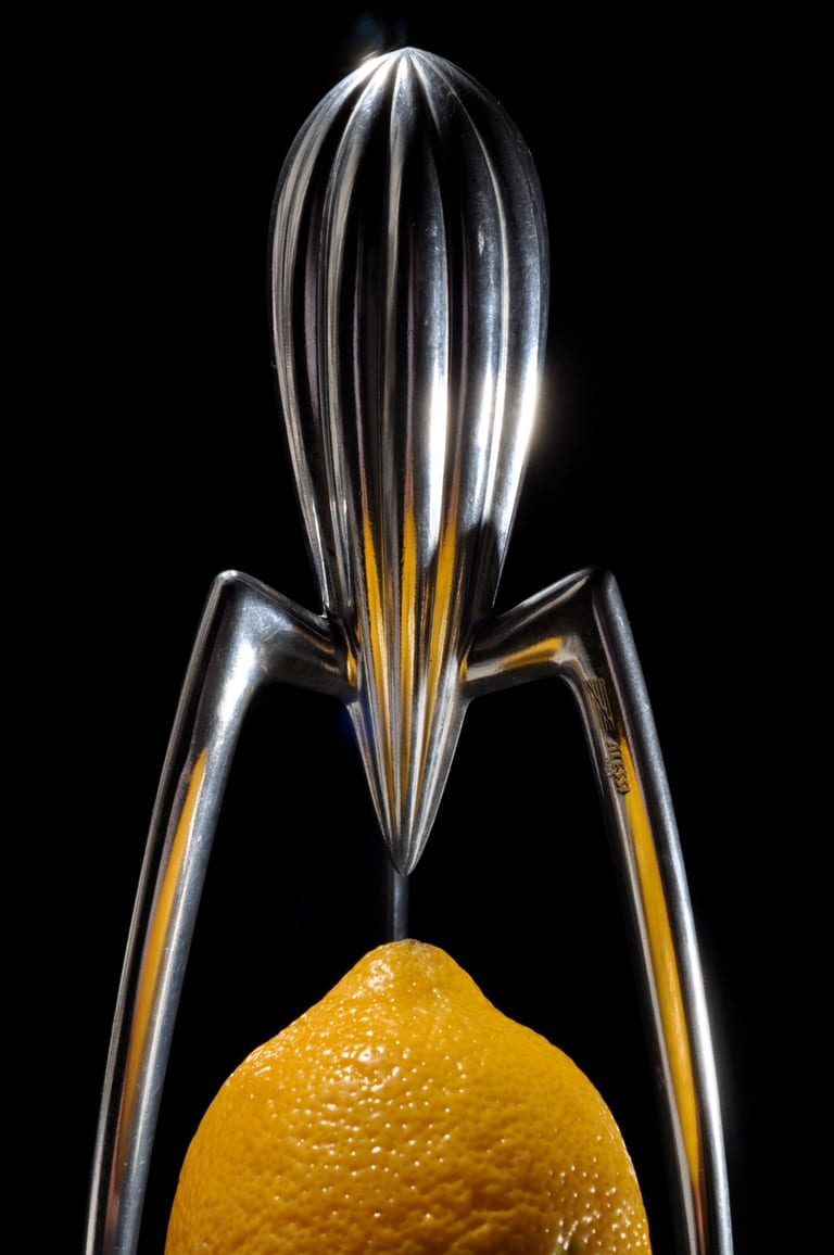 Spegelpolerad Juicy Salif i aluminium med tre långa ben över en citron, mot svart bakgrund
