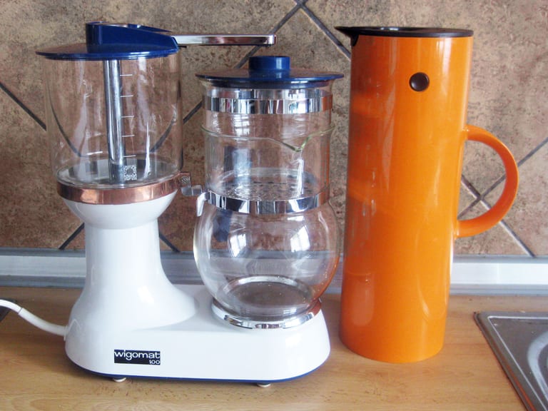 Orange cylindrisk termoskanna från Stelton på en köksbänk bredvid en vit kaffebryggare med glaskannor, mot en kakelvägg