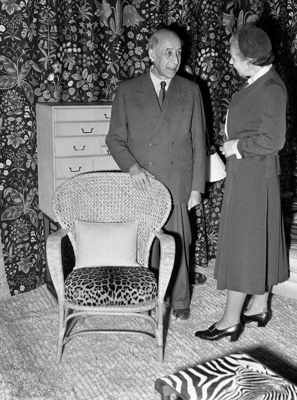 Josef Frank står vid en korgstol med leopardmönstrad dyna och samtalar med Estrid Ericson framför ett blommönstrat tyg på Svenskt Tenns utställning på Nationalmuseum 1952