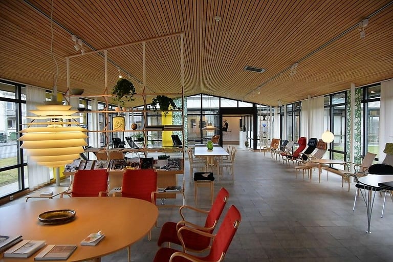 Ljus utställningshall med trätak och glasväggar, möblerad med stolar med röd gjord, bord, en bokhylla och en stor pendellampa
