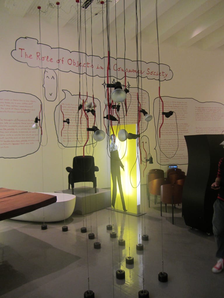 Ett tiotal Parentesi-lampor med runda spotlights som hänger på stålvajrar mellan taket och små svarta vikter på golvet, i en utställning på Triennale Design Museum i Milano