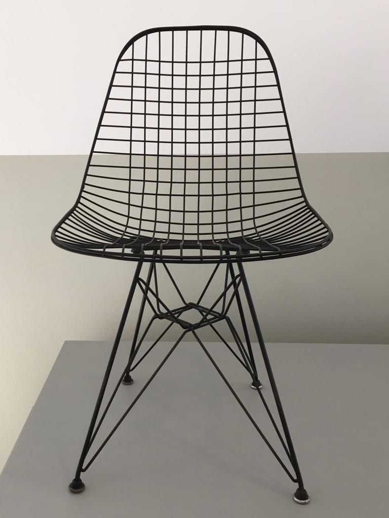 Svart trådstol av Charles och Ray Eames med sits och rygg av svetsat trådgaller på ett underrede av ståltråd, på en grå podie mot en vit och grå vägg