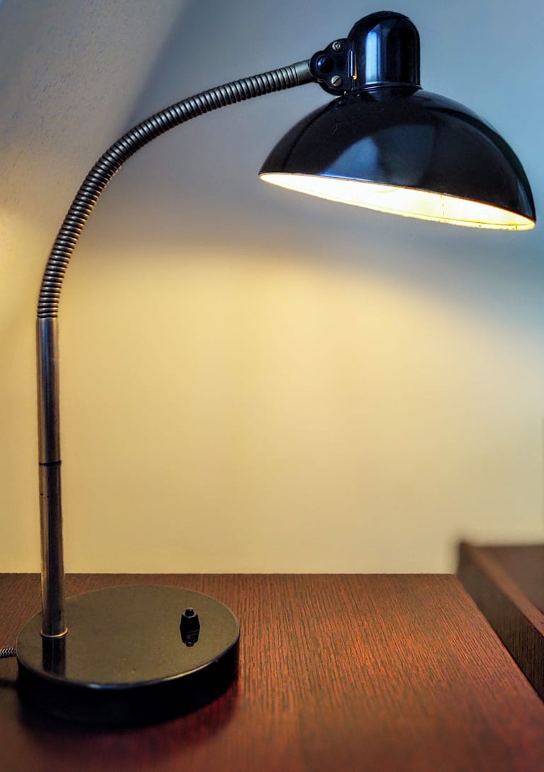 Svart Kaiser idell-skrivbordslampa med böjlig svanhals och kupolformad skärm, tänd, på ett träbord mot en ljus vägg