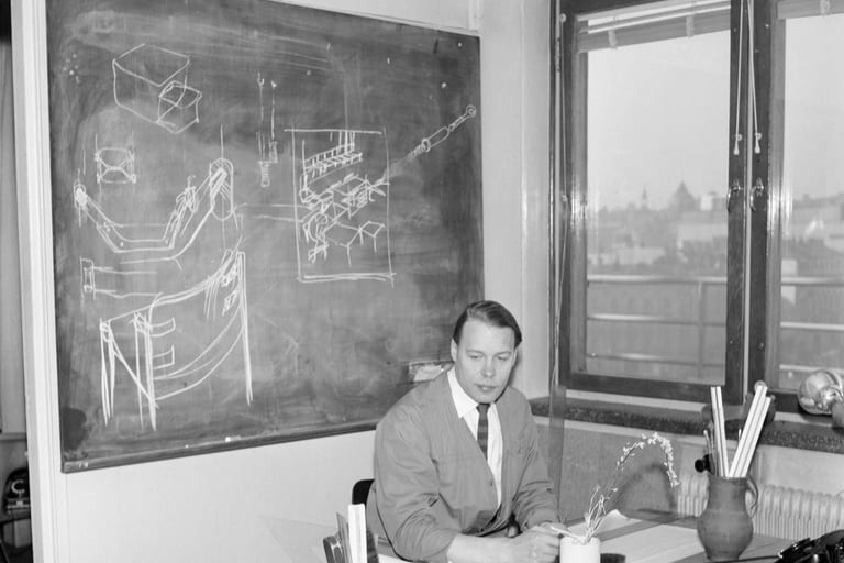 Ilmari Tapiovaara vid sitt skrivbord 1960 framför en svart tavla med kritskisser av möbler och detaljer
