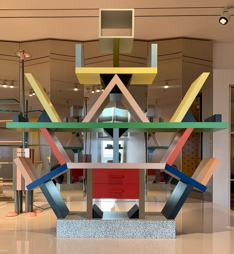 Ettore Sottsass bokhylla Carlton med hyllplan i grönt och gult, diagonala armar i rosa, blått och svart och en bas i svartvitt mönstrat laminat, i ett museirum med speglande väggar