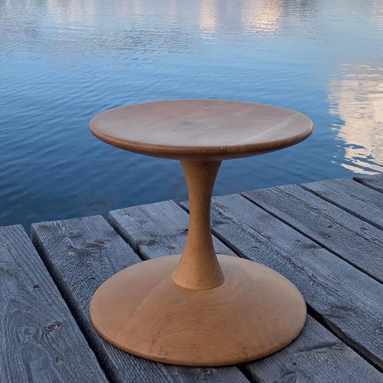 Trisse, en pall i ljust trä med rund sits, smal midja och utsvängd fot, på en träbrygga vid vatten