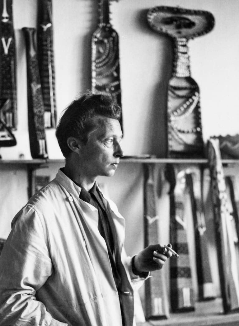 Svartvitt fotografi av Birger Kaipiainen i profil, i ljus arbetsrock och med en cigarett i handen, framför en hylla med långsmala keramikreliefer