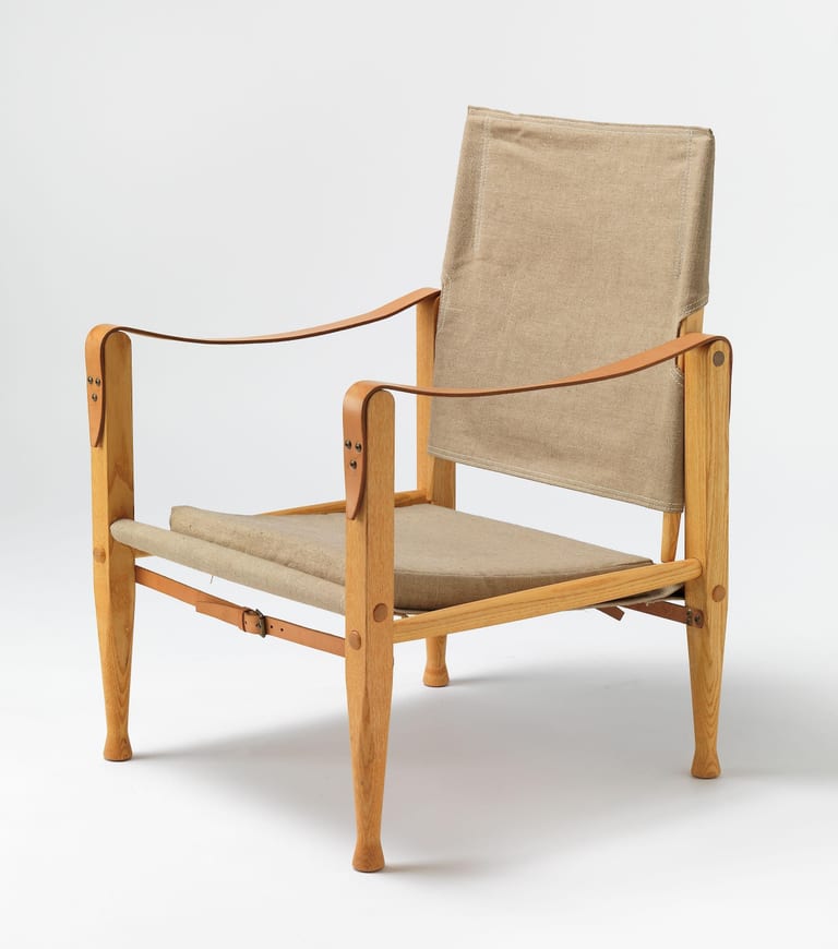Safari Chair i ljus ask med sits och rygg i naturfärgad kanvas, armstöd av läderremmar skruvade i benens överdel och en rem med spänne under sitsen, mot grå bakgrund
