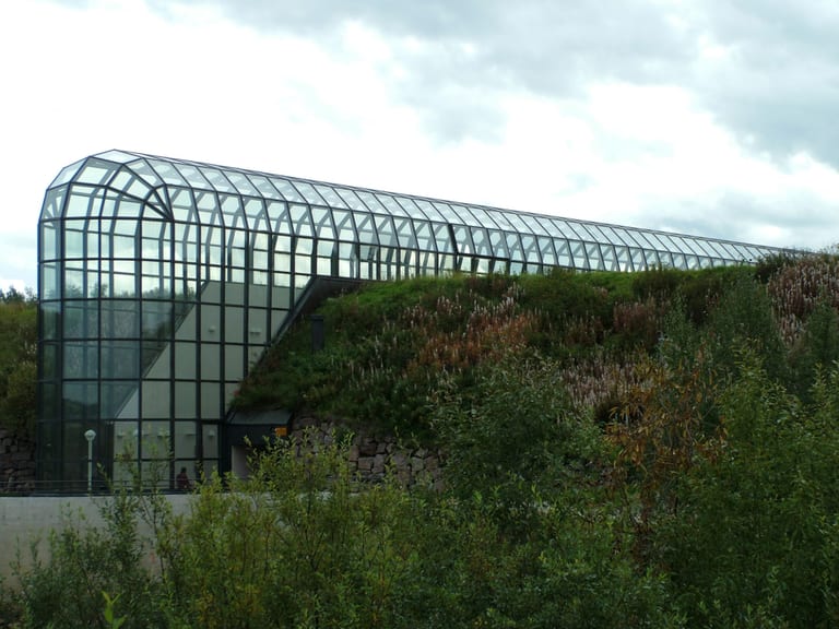 Arktikums långa glasgalleri med välvt tak sträcker sig över en grön, gräsbevuxen kulle, med buskar i förgrunden och mulen himmel