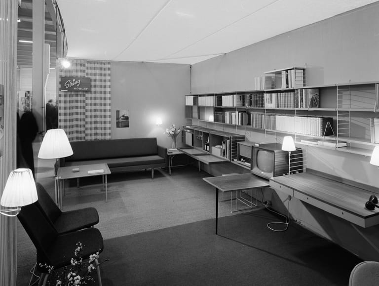 String Furniture – String-hyllor med böcker, radio och tv längs en vägg och en skylt med texten String Möbler, på Sankt Eriksmässan i Stockholm 1957