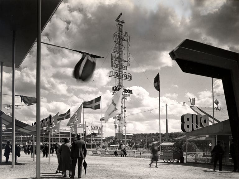 Festplatsen på Stockholmsutställningen 1930 med den höga reklammasten, flaggor, skärmtak och besökare, svartvitt fotografi