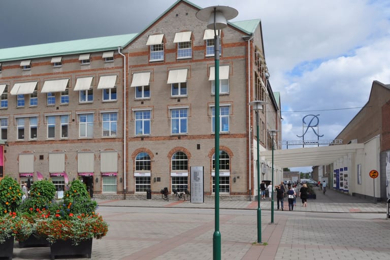 Rörstrand Center i Lidköping: en tegelbyggnad i flera våningar med markiser och grönt plåttak vid ett stenlagt torg, och en portal med Rörstrands R-logotyp