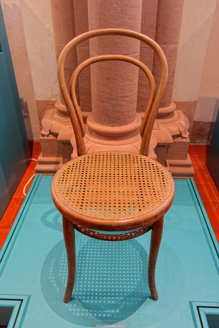 Thonet nr 14 i ljust betsad bok med rund rottingsits, ryggbåge med inre halvbåge och en ring mellan benen, i en museimonter