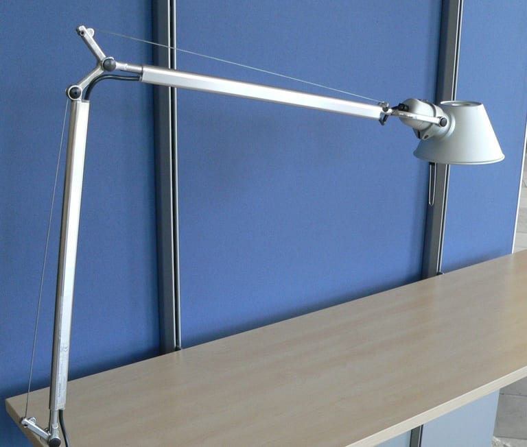 Tolomeo i aluminium fäst vid kanten av en skrivbordsskiva i ljust trä framför en blå skärmvägg, med armen utsträckt och stålvajrar längs båda armdelarna